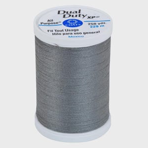 Könnte beinhalten: Eine Spule mit grauem Nähgarn mit weißem Kunststoffober- und -unterteil. Das Etikett trägt die Aufschrift Dual Duty XP, All Purpose, 229 m, Fil Tout Usage, Hilo para uso general und Mexico. Der Faden ist fest um die zylindrische Spule gewickelt.
