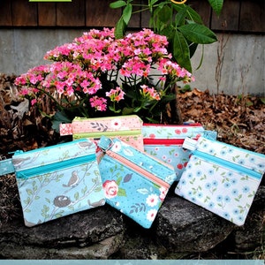 May include: A collection of double zip pouches in various floral and bird-themed fabrics. The pouches have colorful zippers and are arranged on a stone surface, with pink flowers and greenery in the background. The text "Double Zip Pouch" is visible.