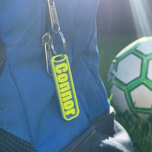 Può includere: Uno zaino blu con cerniera argentata e un portachiavi giallo e verde con la scritta "Connor". Un pallone da calcio è sullo sfondo su un campo erboso.