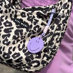Peut inclure: Un sac à bandoulière crème avec un motif léopard noir. Le sac a une fermeture éclair noire et un charm smiley violet attaché. Le texte "VIVI MARI" est visible sur le sac.