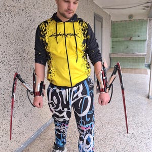 May include: A yellow and black zip-up hoodie with Cyberpunk 2077 text, black joggers featuring white and blue graffiti-style patterns, and two mechanical arm attachments with long, pointed red and silver blades extending from the forearms.