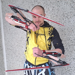 May include: A person holding two red and silver prop blades. The blades are attached to articulated arm pieces with red, black, and silver accents. The person is wearing a yellow and black hoodie and patterned pants.