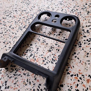 Puede incluir: Un embellecedor de salpicadero de coche de plástico negro con múltiples aberturas para indicadores y controles. El embellecedor tiene un marco rectangular con esquinas redondeadas y una superficie texturizada. Pieza de repuesto.