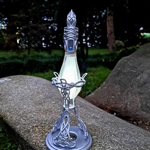 May include: A silver-colored decorative lamp with an intricate design. The lamp has a glowing, light-green glass center and a detailed base with a star-like emblem. The lamp's design is reminiscent of fantasy or sci-fi themes.
