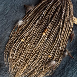 May include: Handmade dreadlocks in shades of brown and blonde, adorned with white thread accents, wooden beads, and feathers. The dreadlocks are arranged on a wooden stick, creating a bohemian and natural aesthetic.