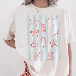 Puede incluir: Camiseta color crema con diseño náutico. La camiseta tiene rayas verticales azul claro con ilustraciones rojas de conchas marinas, estrellas de mar y un caballito de mar. La camiseta es de corte holgado.