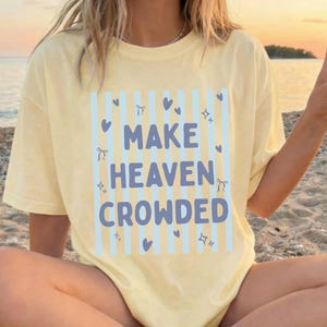 Puede incluir: Una camiseta de manga corta de color amarillo pálido con un fondo de rayas verticales azul claro, que presenta el texto "MAKE HEAVEN CROWDED" en letras azules negritas, decorado con pequeños iconos de corazones y estrellas azules.