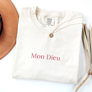 May include: A cream-colored short-sleeve t-shirt featuring the text Mon Dieu embroidered in red thread on the chest. The shirt has a crew neckline with a Comfort Colors brand label inside the collar and is paired with a gold-toned pendant necklace.