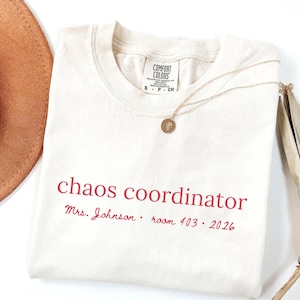Puede incluir: Una camiseta de color crema con el texto en rojo "chaos coordinator" seguido de "Mrs. Johnson • room 403 • 2026". La camiseta presenta una etiqueta de la marca Comfort Colors en el cuello y se muestra con un collar de colgante circular dorado.