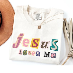 Puede incluir: Camiseta color crema con la frase "Jesus Loves Me" en un estilo recortado y colorido. La camiseta tiene cuello redondo y mangas cortas. Un collar de oro con un colgante está drapeado sobre la camiseta. Un sombrero marrón y una planta seca están en el fondo.