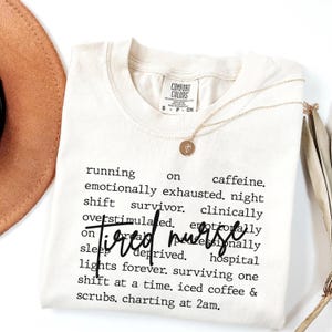 Puede incluir: Camiseta color crema con las palabras "Tired Nurse" en fuente negra. La camiseta tiene una lista de frases en una fuente más pequeña. Un collar dorado con un colgante está sobre la camiseta.