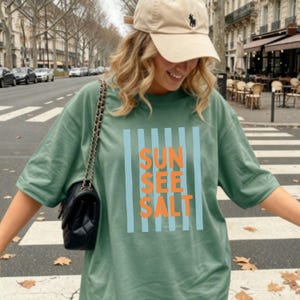 May include: Sage green t-shirt with the words "SUN SEE SALT" in orange letters, framed by light blue vertical stripes. The shirt is paired with a tan baseball cap and a black shoulder bag. The image is taken outdoors.
