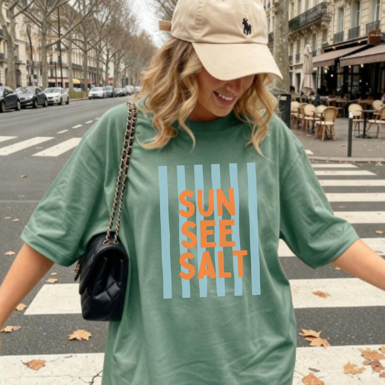 May include: Sage green t-shirt with the words "SUN SEE SALT" in orange letters, framed by light blue vertical stripes. The shirt is paired with a tan baseball cap and a black shoulder bag. The image is taken outdoors.
