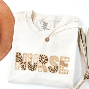 Puede incluir: Camiseta color crema con la palabra "NURSE" en letras de patchwork. Las letras presentan estampados de leopardo, cuadros y lunares en tonos marrón y beige. Un collar dorado con un colgante descansa sobre la camiseta.