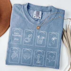 Puede incluir: Una camiseta azul Comfort Colors con un diseño gráfico blanco. El diseño incluye el texto "Kate's bachelorette 2025" e ilustraciones de un anillo, un cóctel, una palmera, una tortuga, una concha, un letrero de playa y una estrella de mar. Un collar dorado está sobre la camiseta.