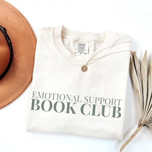 Puede incluir: Camiseta color crema con el texto "EMOTIONAL SUPPORT BOOK CLUB" en verde. Un collar dorado con un colgante y un sombrero marrón están junto a la camiseta. Una hoja de palma seca también está en la imagen.