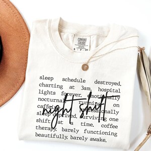 Puede incluir: Camiseta Comfort Colors color crema con las palabras "night shift" en negrita y cursiva. La camiseta presenta una lista de palabras relacionadas con los turnos de noche en negro, con fuente sans-serif. Un collar dorado con un colgante está sobre la camiseta.