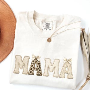 Puede incluir: Camiseta color crema con la palabra "MAMA" en una fuente decorativa. La letra "M" y la última "A" tienen lazos, y la "A" del medio tiene un diseño de estampado de leopardo. También se ve un collar dorado con un colgante.