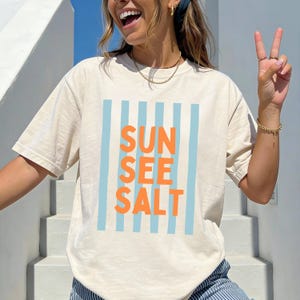 May include: Cream-colored t-shirt with the words "SUN SEE SALT" in orange, set against a backdrop of light blue vertical stripes. The shirt has a relaxed fit and short sleeves, perfect for casual wear.