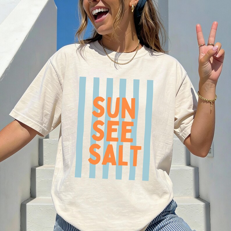 May include: Cream-colored t-shirt with the words "SUN SEE SALT" in orange, set against a backdrop of light blue vertical stripes. The shirt has a relaxed fit and short sleeves, perfect for casual wear.