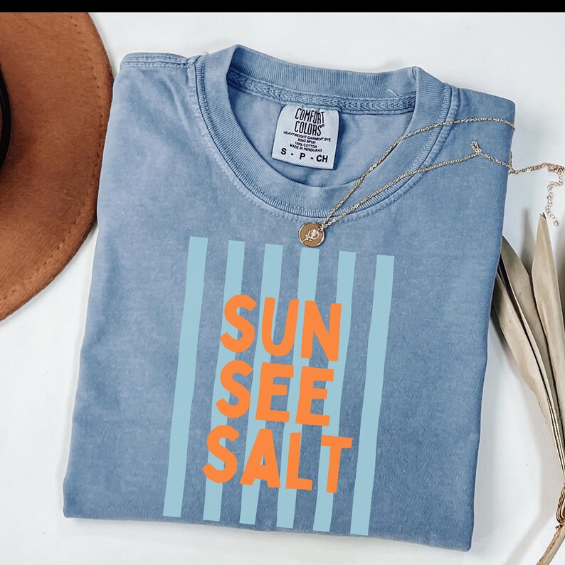 May include: A blue Comfort Colors t-shirt with the words "SUN SEE SALT" in orange, framed by vertical light blue stripes. A gold necklace with a pendant rests on the shirt. A brown hat is partially visible.