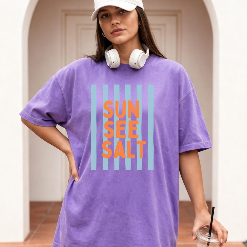 May include: A purple t-shirt with the words "SUN SEE SALT" in orange letters, framed by vertical light blue stripes. The shirt has short sleeves and a relaxed fit. The model is wearing a white cap and headphones.