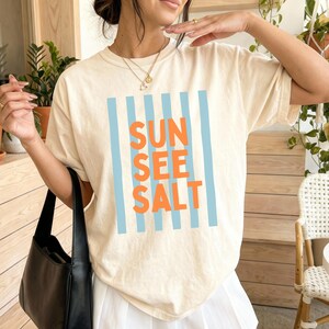 May include: Cream-colored t-shirt with the words "SUN SEE SALT" in orange block letters, framed by vertical light blue stripes. The shirt has a relaxed fit and short sleeves, perfect for casual wear.