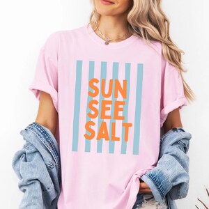 May include: A light pink t-shirt with the words "SUN SEE SALT" in orange block letters, framed by light blue vertical stripes. The shirt has rolled-up sleeves and is part of a casual summer clothing collection.