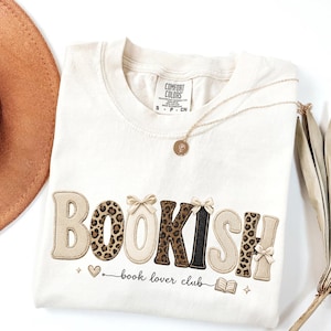 Puede incluir: Camiseta color crema con la palabra "BOOKISH" en una fuente decorativa, con estampado de leopardo y lazos. La frase "book lover club" está impresa debajo. También se ven un collar dorado y un sombrero marrón.