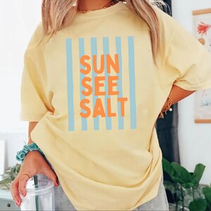 May include: A yellow t-shirt with the words "SUN SEE SALT" in orange letters, framed by vertical light blue stripes. The shirt is a casual, oversized fit.