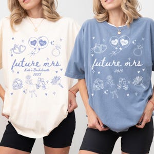 Puede incluir: Dos camisetas, una crema y otra azul, con el texto "future mrs 2025" en letra cursiva. Las camisetas tienen un gráfico de globos, corazones, un anillo, pastel, flores, copas de champán y un ramo. La camiseta crema dice "Kate's Bachelorette".
