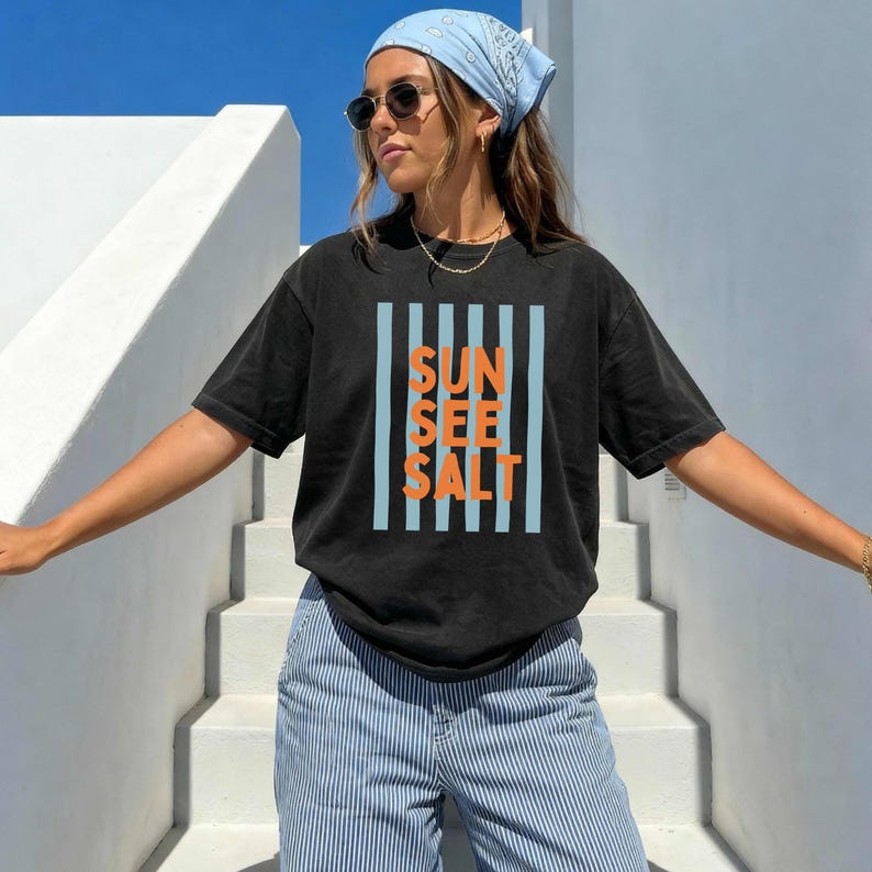 May include: Black t-shirt with the words "SUN SEE SALT" in orange letters, framed by vertical light blue stripes. The shirt is worn with blue and white striped pants and a light blue bandana.