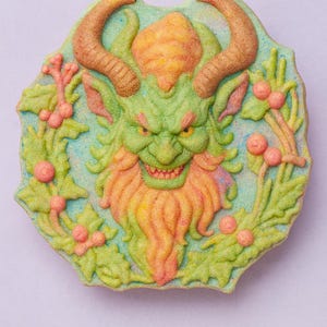 以下が含まれることがあります： 角のある生き物の顔の形をしたデコレーションクッキー。緑色の顔、オレンジ色のひげと角、水色の背景が特徴です。クッキーは緑の葉とピンク色のベリーに囲まれています。