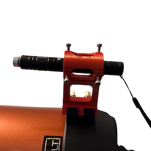 May include: A red 3D printed laser mount with a black laser pointer attached to a telescope. The mount has two silver screws on each side and a black strap.