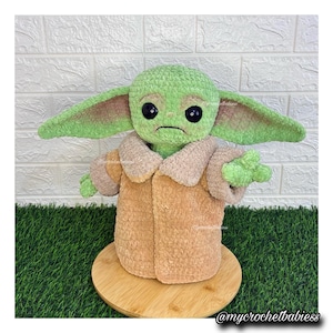 May include: A handmade crochet doll of a character with large ears, green skin, and a tan coat. The doll has large black eyes and a slightly sad expression. The doll is sitting on a wooden base, with a green and white background. The text "@mycrochetbabiess" is visible.