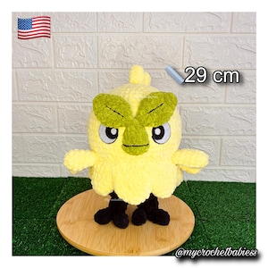 May include: A handmade, yellow crochet bird plush toy, approximately 29 cm tall. The bird has green leaf-shaped accents, black feet, and large, expressive eyes. The toy is displayed on a wooden surface.