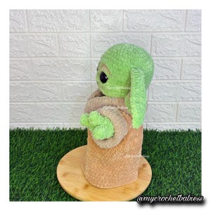 May include: A handmade green and tan crochet doll, resembling a popular character. The doll has large black eyes and long, floppy ears. It is wrapped in a tan-colored blanket and sits on a wooden base. The doll is a soft, plush toy.