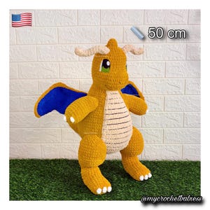 May include: A handmade crochet Dragonite plush toy, measuring approximately 50 cm in height. The toy features a golden-yellow body, blue wings, and a cream-colored belly. It has green eyes and white accents. The toy is displayed on a green surface.