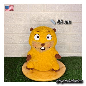May include: A handmade, yellow crochet groundhog plush toy, approximately 10 inches tall, with brown accents on its ears, paws, and nose. The groundhog has large, white-rimmed eyes and a smiling mouth. It sits on a wooden round base.