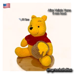 Op de afbeelding: Een handgemaakt gele Winnie de Poeh pluche speelgoed, ongeveer 28 cm hoog, met een rood shirt en een bruine honingpot met gele honing. Gemaakt met Alize Velluto garens en een 6 mm haak.