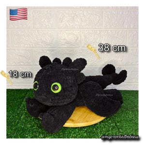 May include: A black crochet dragon plush toy, 18 cm tall and 38 cm long, with large green eyes. The plush is lying on a wooden surface against a white brick wall and green grass. The American flag is in the top left corner.