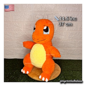 May include: An orange crochet Charmander plush toy, 14.5 inches tall, with a yellow belly and white accents on its feet. The toy is displayed on a wooden stand against a textured white brick wall.