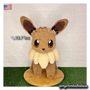 Peut inclure: Une peluche Eevee faite à la main, d'environ 34,8 cm de haut, dans des tons de marron et de crème. L'animal en peluche a de grands yeux expressifs et un aspect duveteux et texturé. Il est présenté sur une base en bois.