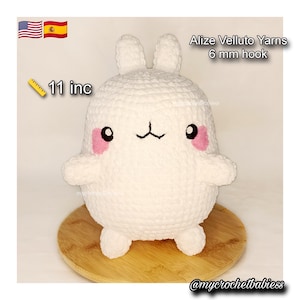 May include: A white, plush, crocheted bunny toy with pink cheeks and a black facial expression. The toy is approximately 11 inches tall and made with Alize Velluto Yarns using a 6 mm hook. The toy is sitting on a wooden surface.