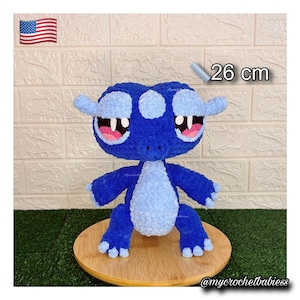 Può includere: Un giocattolo a forma di creatura all'uncinetto blu e azzurro fatto a mano, alto circa 26 cm. Il giocattolo ha grandi occhi con accenti rosa ed è esposto su una superficie di legno. Il testo "26 cm" è visibile.