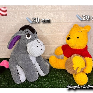 Puede incluir: Dos peluches de ganchillo: un burro gris con orejas moradas y un oso amarillo con una camisa roja. El burro mide 28 cm de alto. El oso sostiene un tarro de miel. Los juguetes están sobre hierba verde.