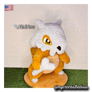 Peut inclure: Une peluche Cubone faite à la main au crochet, d'environ 31,75 cm de haut. La peluche présente une tête en forme de crâne blanche, des yeux orange et un corps beige et orange. Elle tient un os et est présentée sur une base en bois.