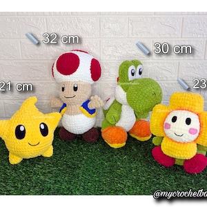 Könnte beinhalten: Vier gehäkelte Plüschtiere: ein gelber Stern (21 cm), eine Pilzfigur (32 cm), ein grüner Dinosaurier (30 cm) und eine Blumenfigur (23 cm). Die Spielzeuge befinden sich auf einer grünen Oberfläche mit einem weißen Ziegelhintergrund. Die Spielzeuge sind handgefertigt.