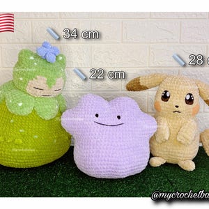 Op de afbeelding: Drie gehaakte pluche speelgoed: een groen-witte figuur (34 cm), een lavendelkleurige Ditto (22 cm) en een gele Pikachu (28 cm). Het speelgoed wordt weergegeven op een groene ondergrond met een witte bakstenen muur als achtergrond. De Amerikaanse vlag staat linksboven.