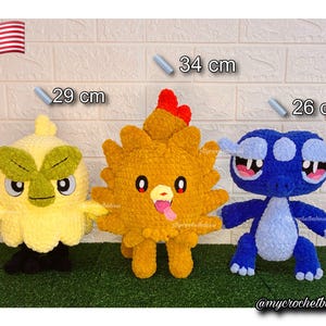 May include: Three handmade crochet plush toys. A yellow bird-like creature is 29 cm tall, a golden chicken-like creature is 34 cm tall, and a blue creature is 26 cm tall. The toys are on a green surface.